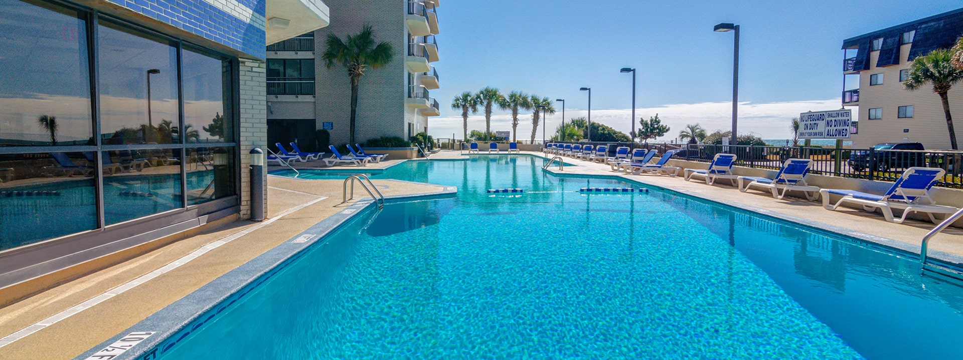 A large outdoor pool with blue water and lounge chairs, surrounded by palm trees and a beige building.