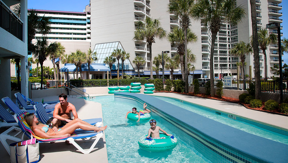 A family enjoys a sunny day by the pool with inflatable toys and lounge chairs nearby.
