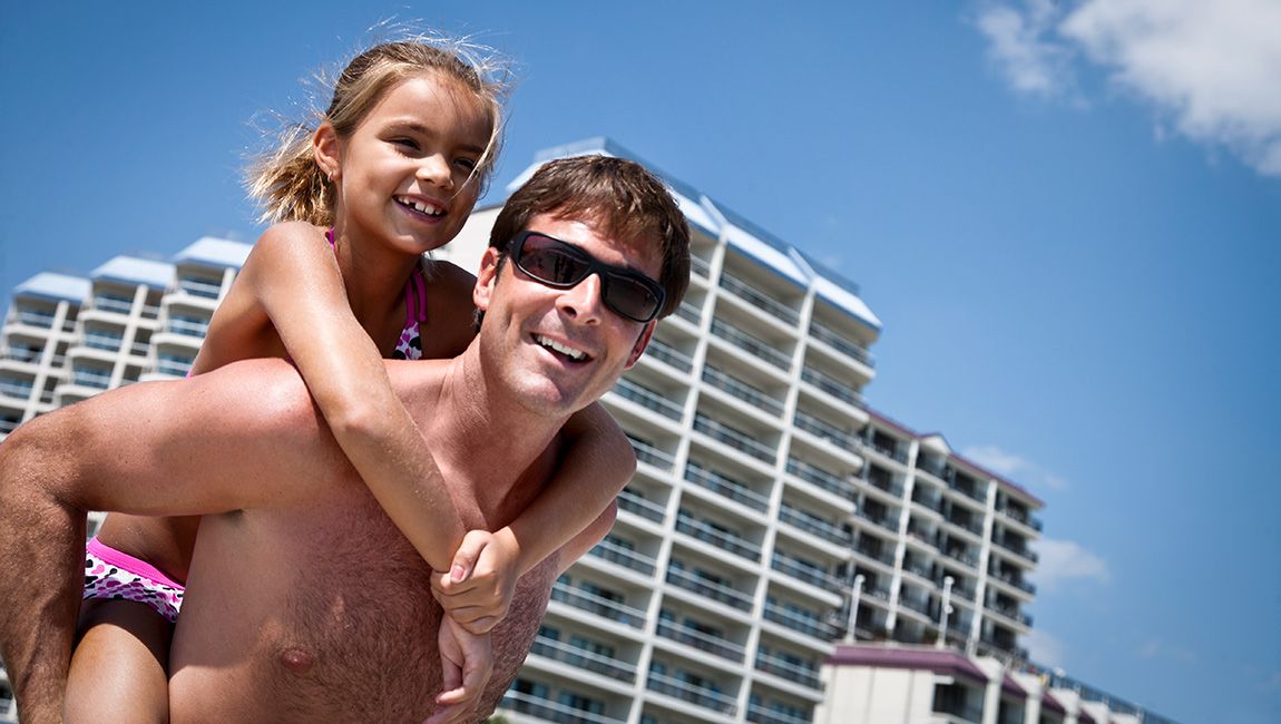 A man carrying a young girl on his back in front of a large building with balconies.