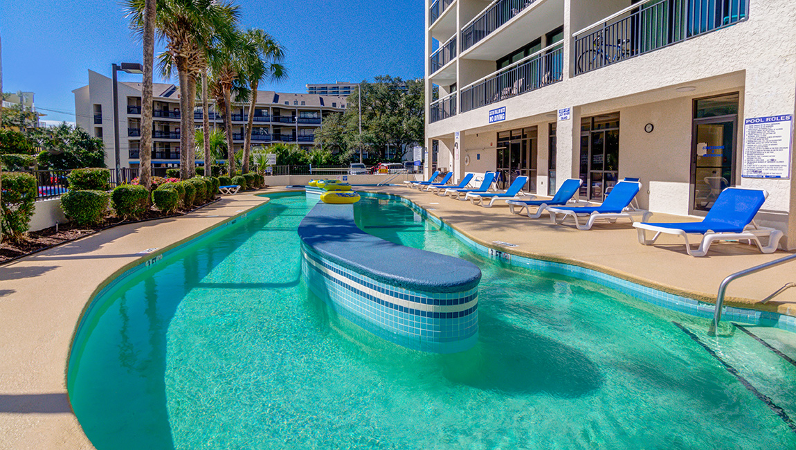A swimming pool with a curved bar and blue lounge chairs, surrounded by palm trees and a building with balconies.