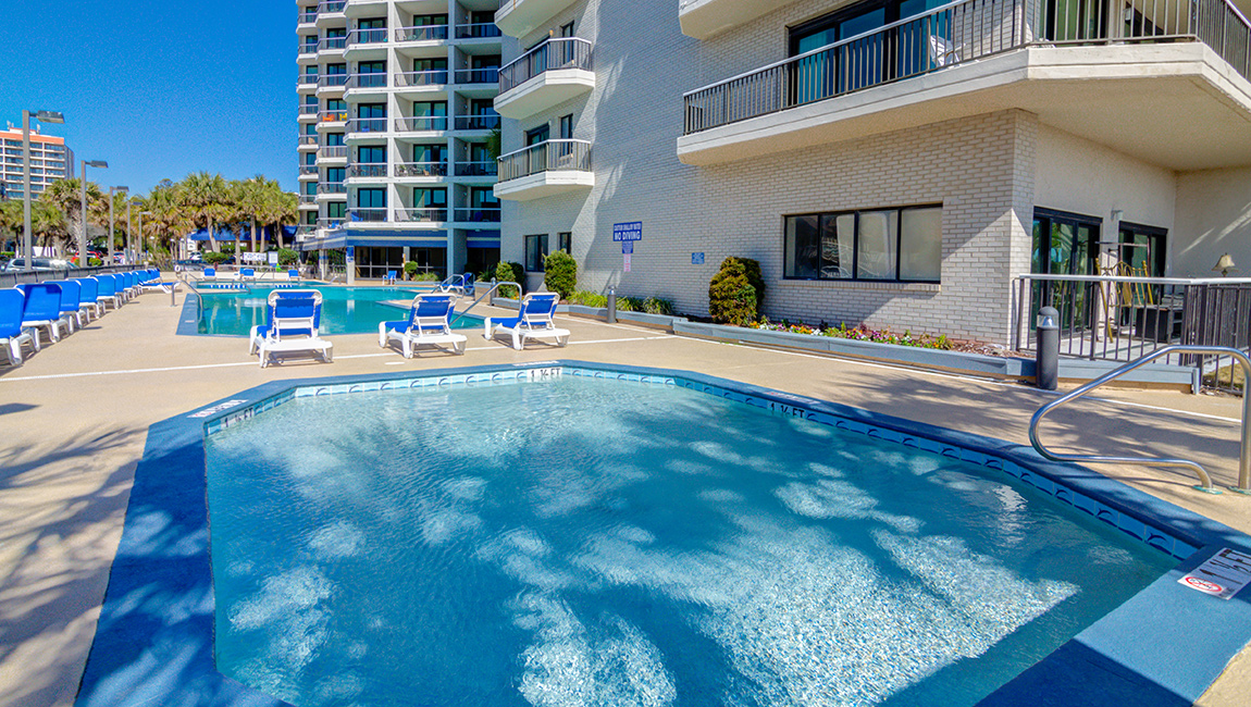 A swimming pool with blue water and white lounge chairs next to a tan building with balconies.